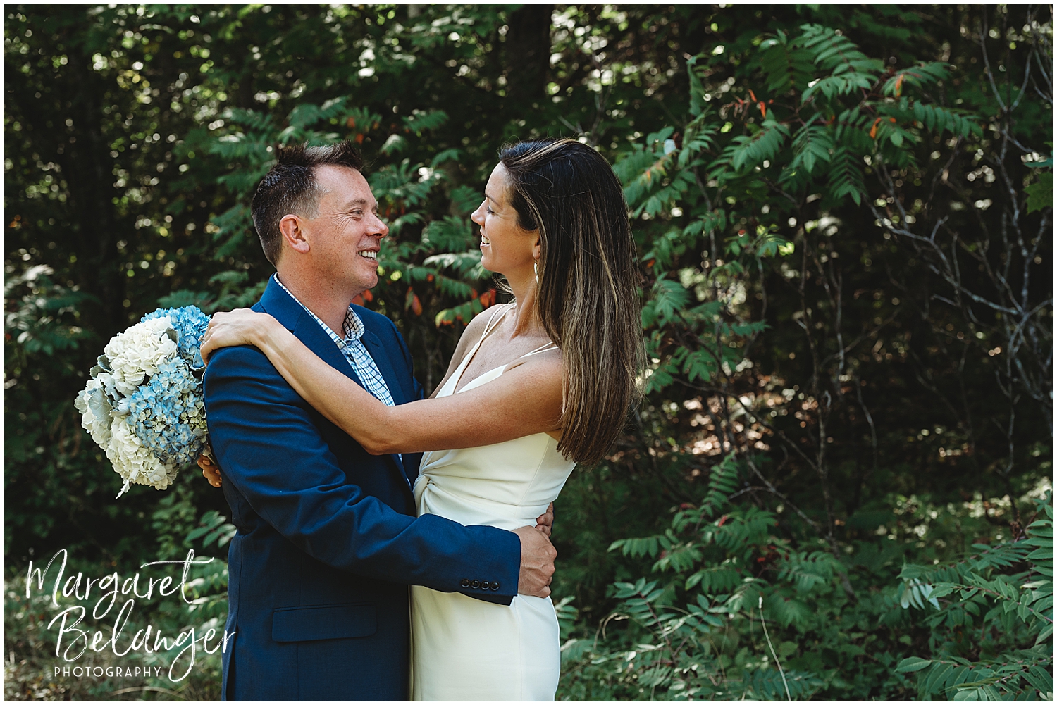A happy couple embracing in a wooded area, with the bride on the right holding a bouquet of blue and white hydrangeas.