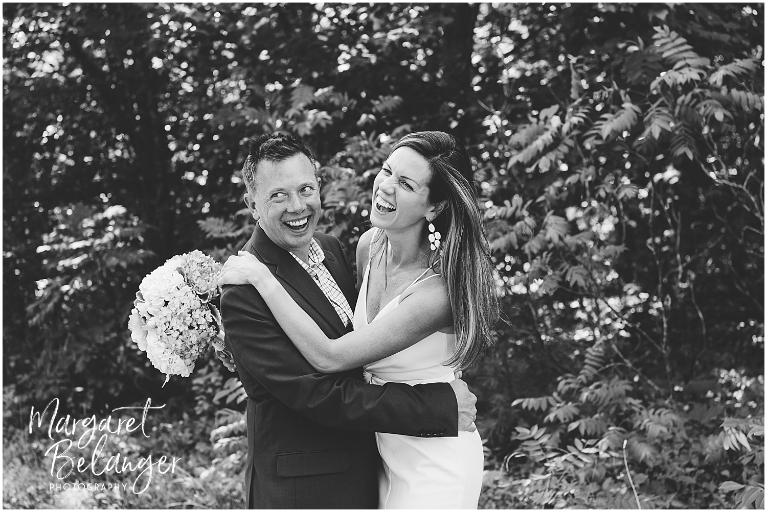 A happy wedding couple laughing together while posing for a photo, the bride holding a bouquet of flowers.