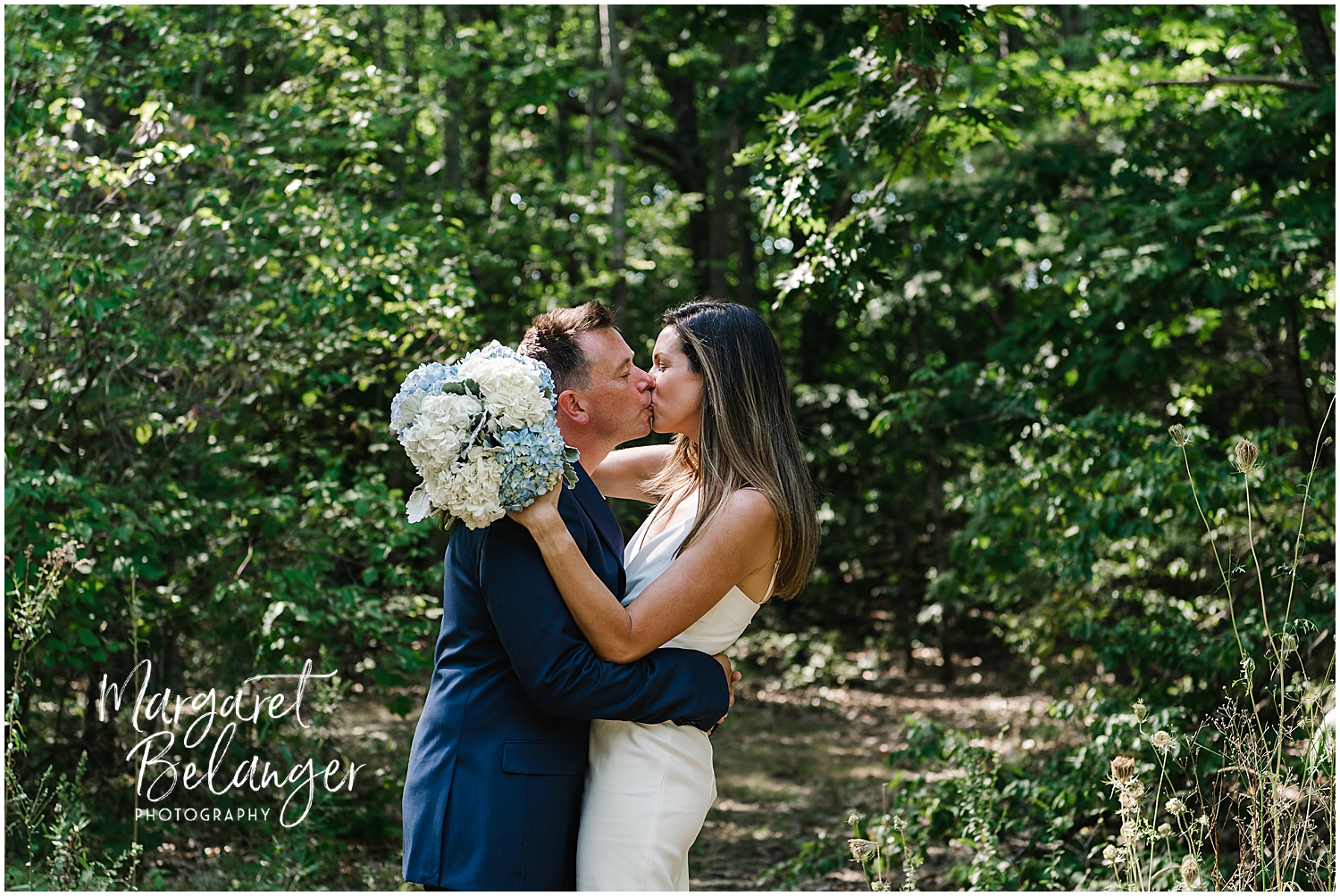 A couple kissing amidst greenery with the woman holding a bouquet of hydrangeas.