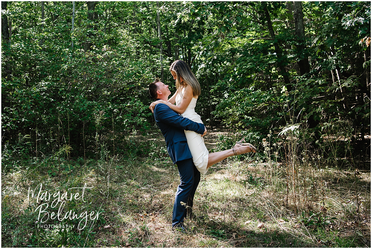 A groom in a blue suit lifting a bride in a simple white wedding dress in a forest clearing.