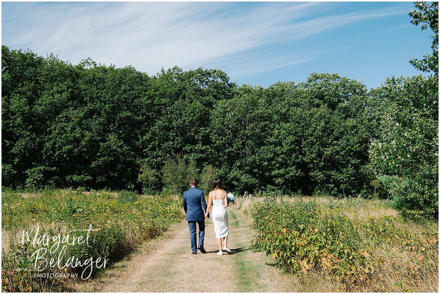 A bride and groom holding hands and walking down a natural path flanked by lush greenery under a clear blue sky.