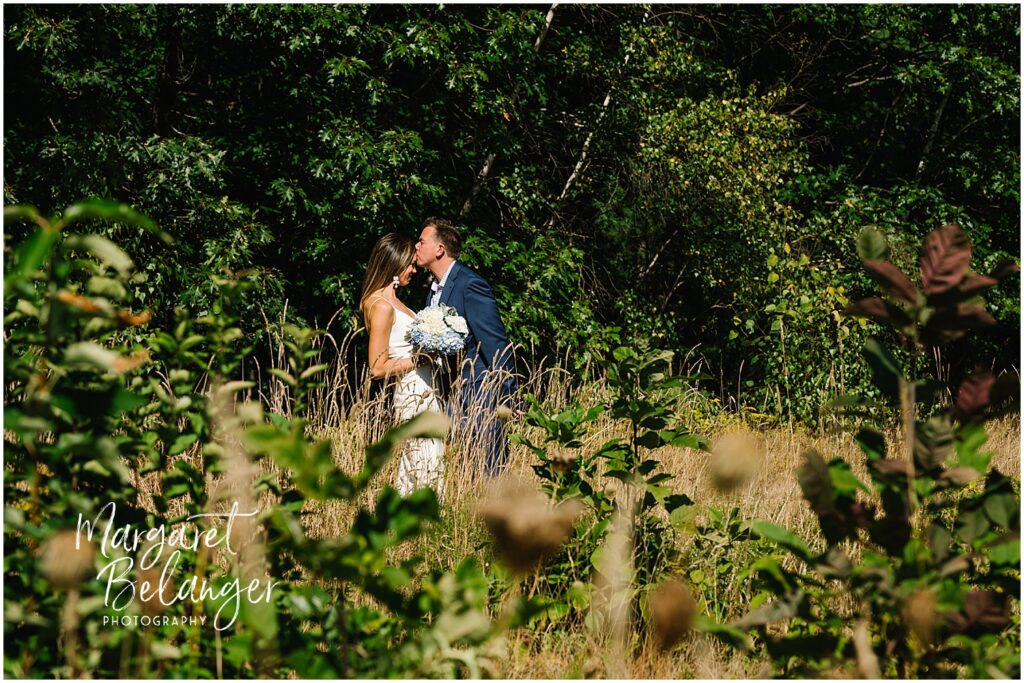 A groom kisses his bride's forehead amidst tall grass and greenery.