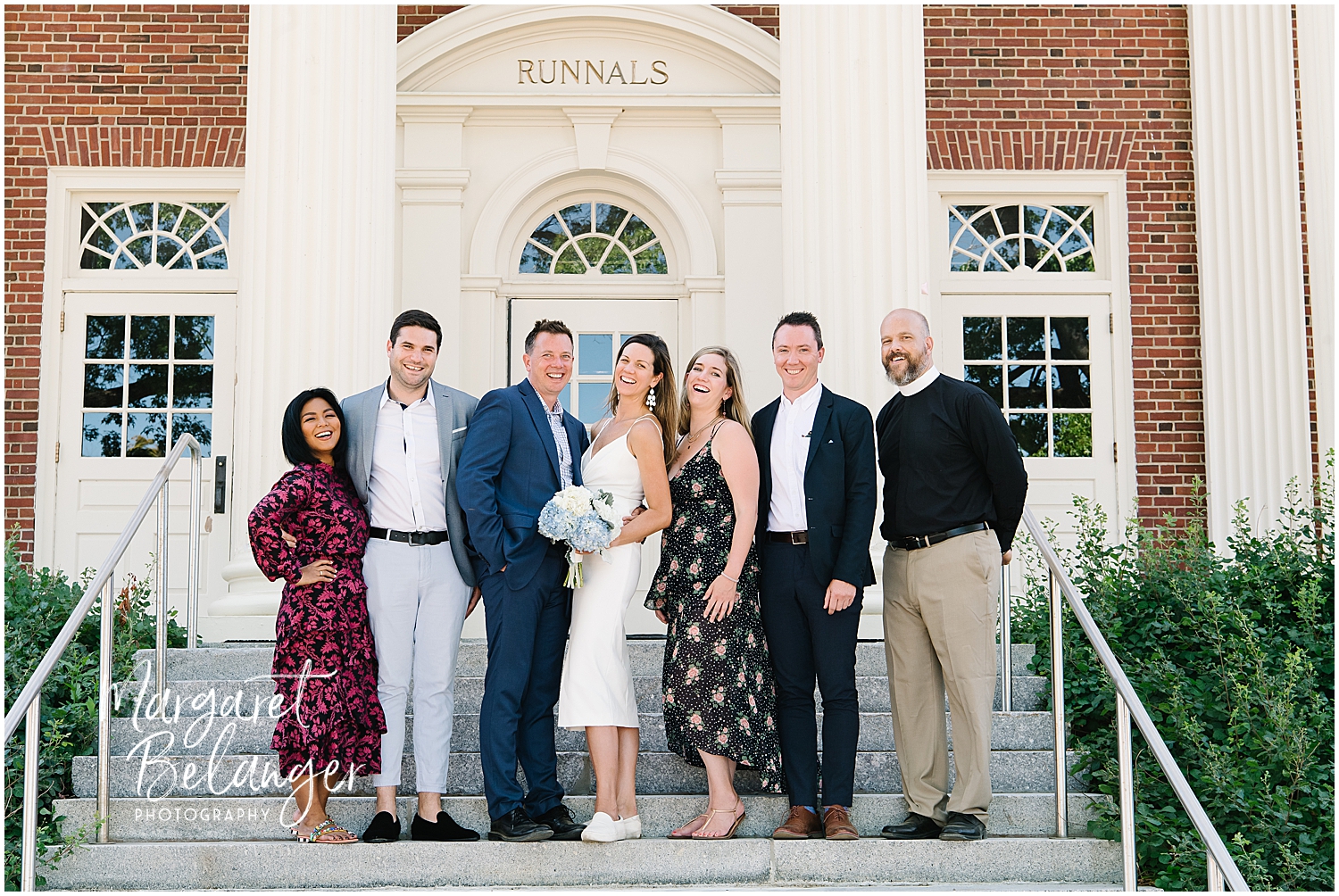 A group of well-dressed individuals smiling and posing together on the steps of a building with the word "Runnals" engraved above the entrance.