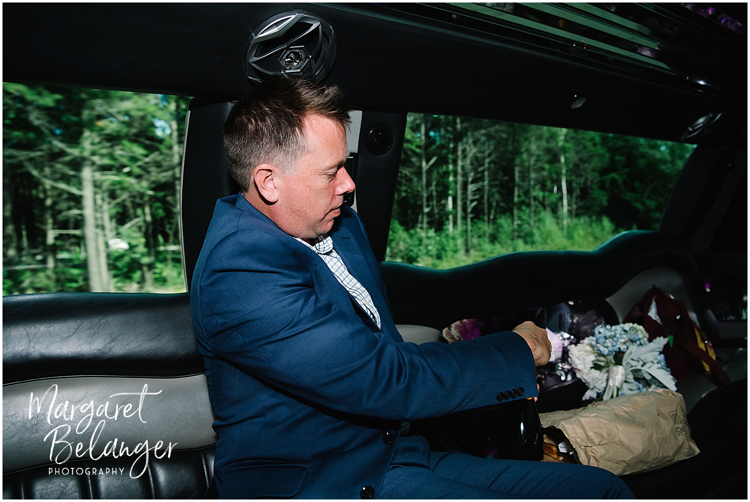Groom in a suit popping champagne in a limousine with a bouquet of flowers on the seat next to him.