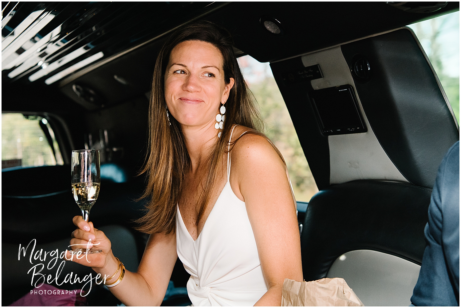 A bride holding a champagne flute while sitting inside a limousine, smirking at the groom who is off-camera.