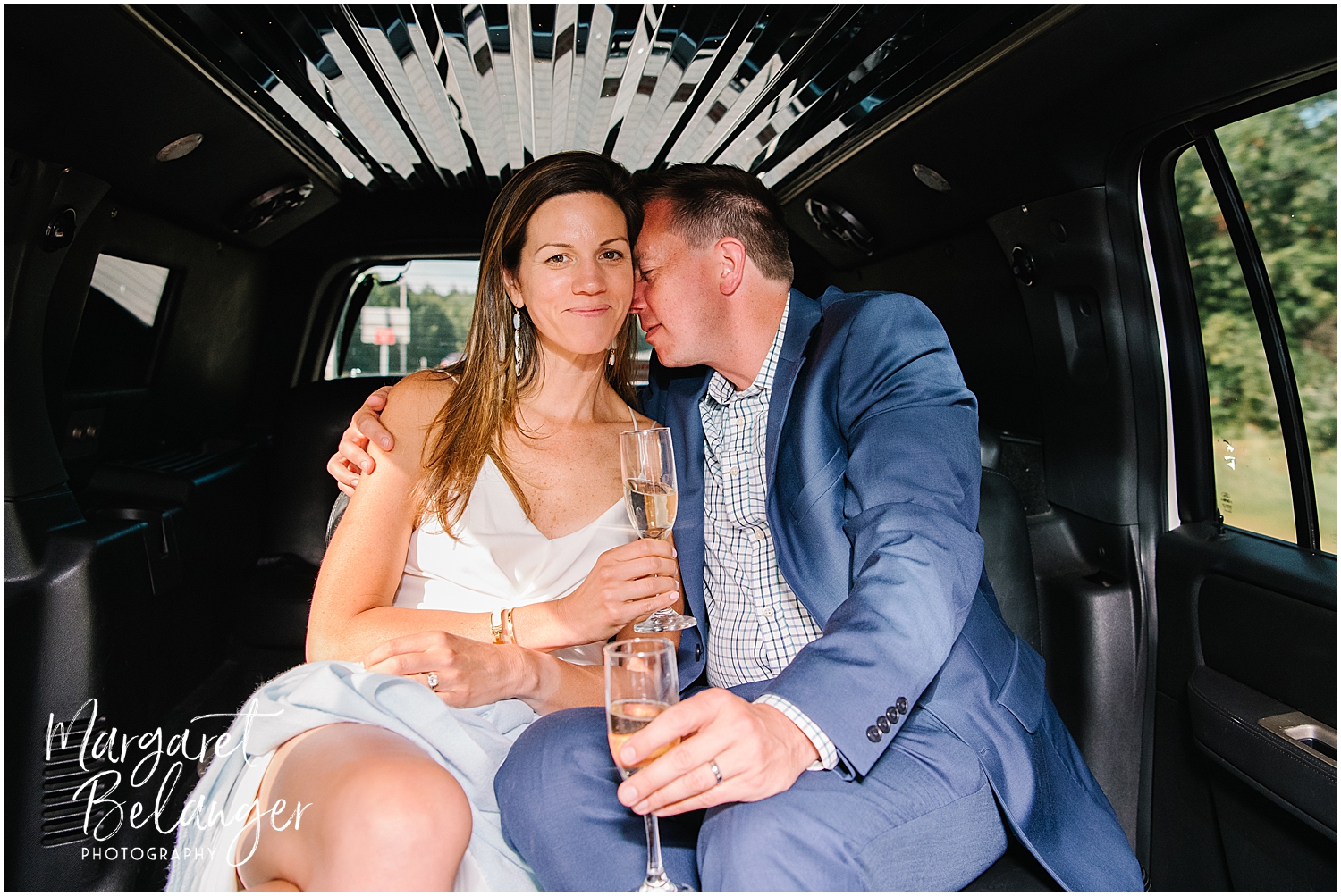 The groom nuzzles the bride in the back of the limousine while they hold flutes of champagne.