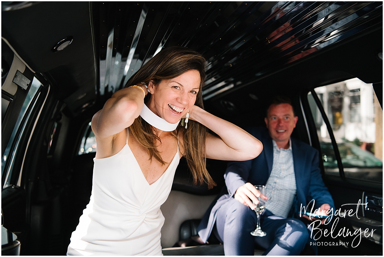 A bride laughs while adjusting a clerical collar inside a limousine with the groom in a suit seated in the background.