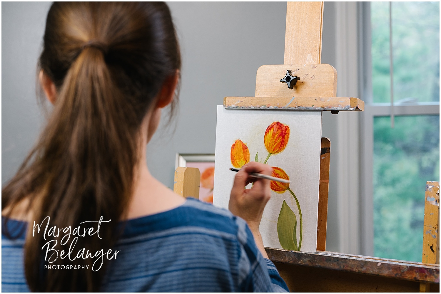 A person with brown hair paints orange tulips on a canvas placed on an easel, with a window in the background.