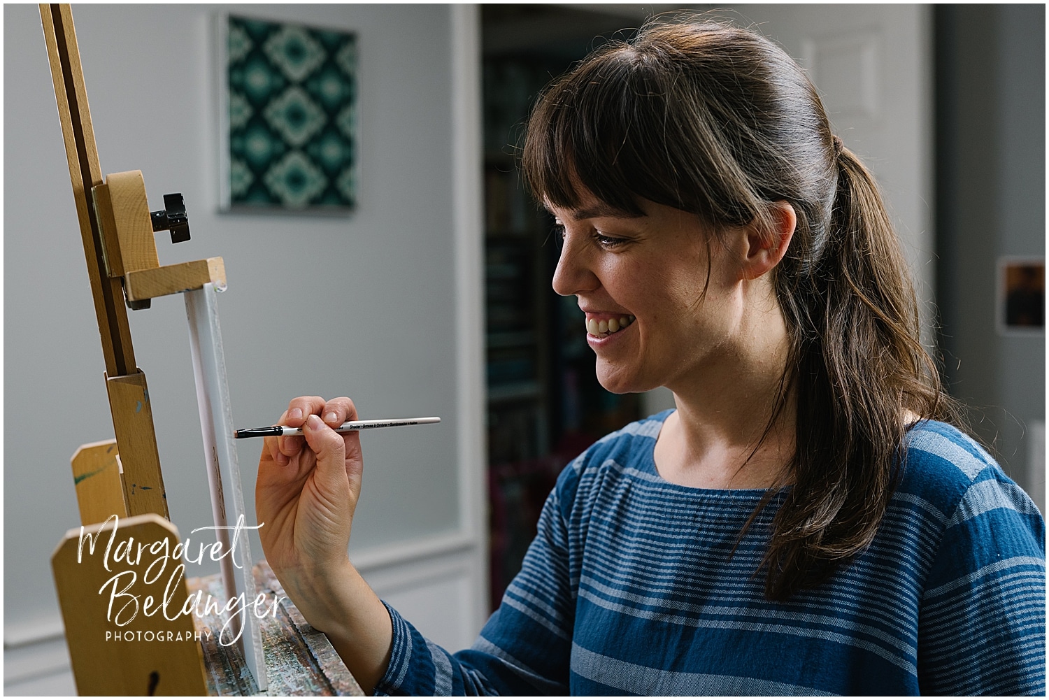 A smiling woman with a ponytail, wearing a blue-striped shirt, paints on a canvas positioned on an easel inside a room.