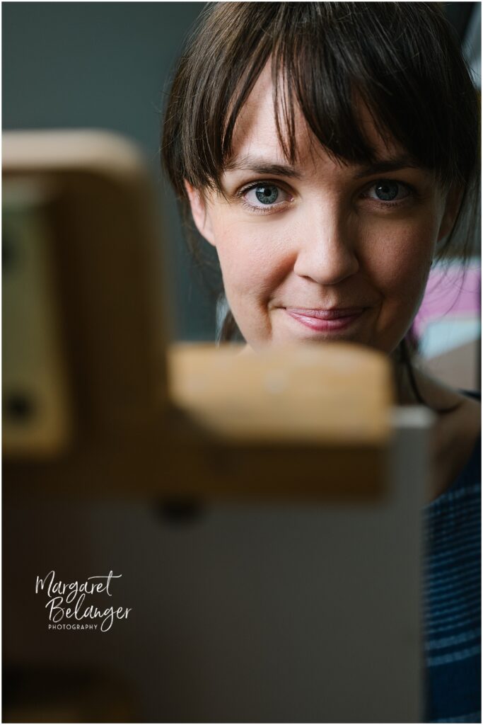 A woman with bangs and blue eyes looks towards the camera, partially obscured by her easel.